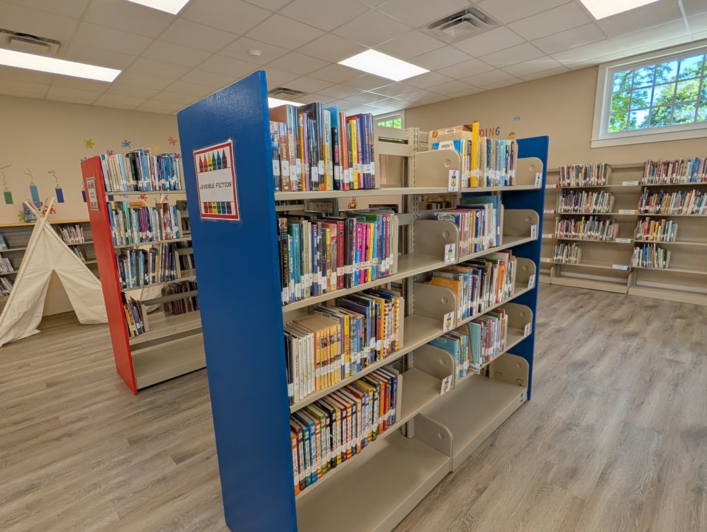 Bookshelves filled with juvenile fiction books in a brightly lit library, with a reading teepee and more bookshelves in the background.
