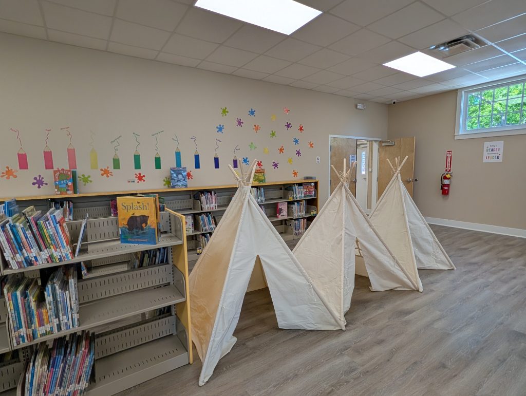 Children's library area with bookshelves, colorful wall decorations, and three small fabric teepees arranged on a wooden floor near a window.