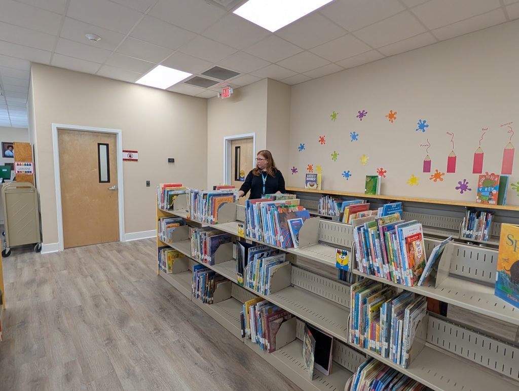 Woman stands behind bookshelves with children’s books in a library, colorful crayon and splatter wall decorations in the background, and closed wooden doors visible.