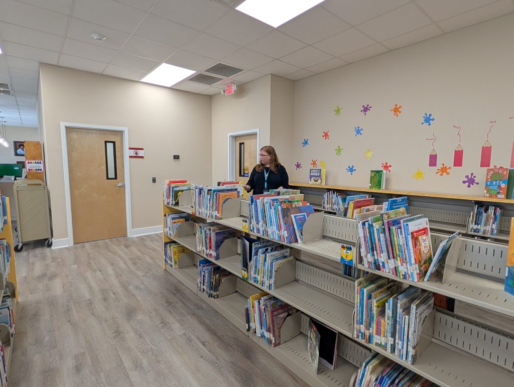 A woman stands in a children's library section with bookshelves, colorful wall decorations, and wooden doors.