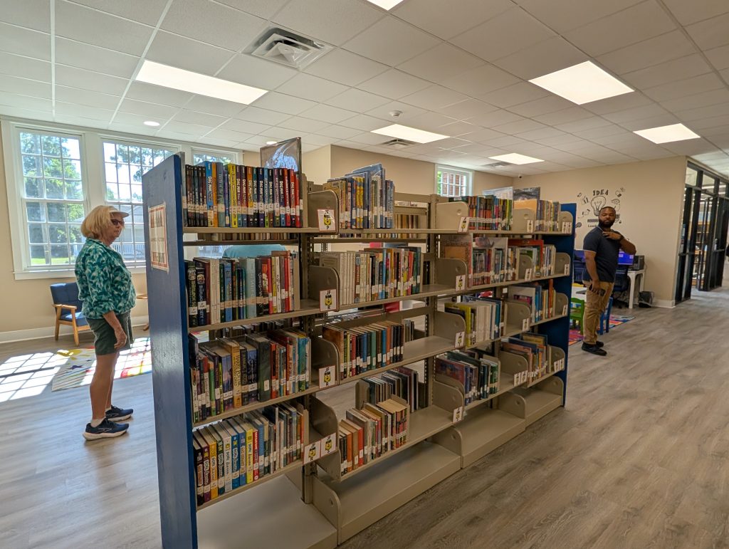 Two people stand in a brightly lit library, browsing bookshelves filled with a variety of books. Large windows let in natural light.