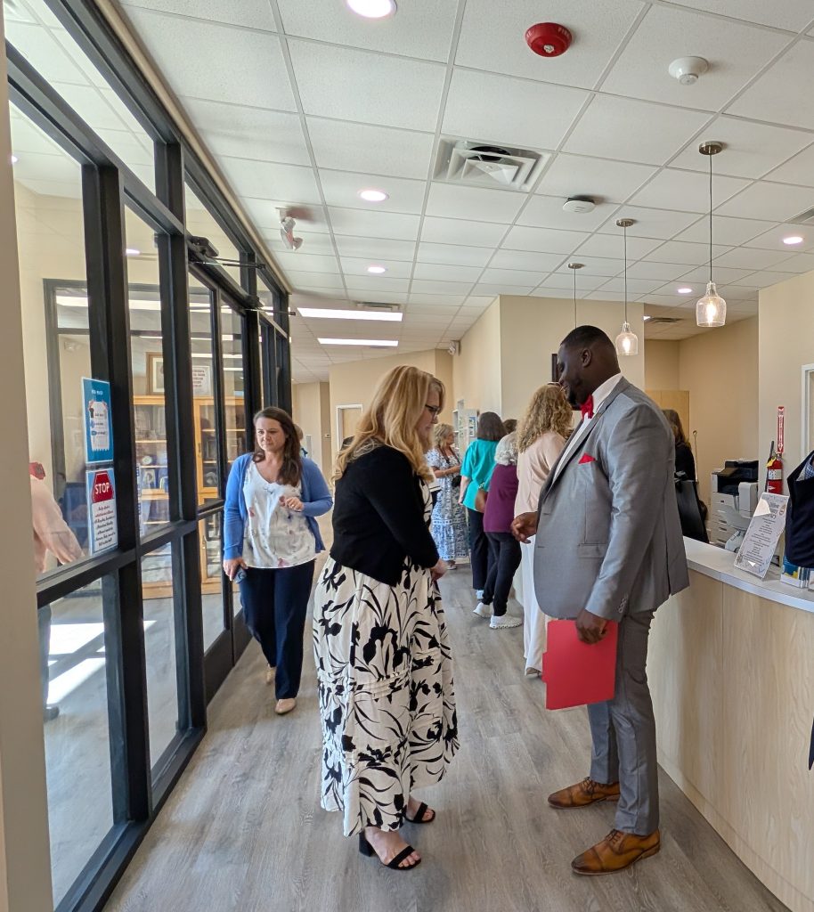 People conversing in a bright office lobby with large windows, modern lighting, and a reception desk. Some individuals walk in the background.