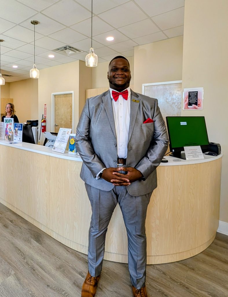 Man standing in front of a reception desk, dressed in a gray suit with a red bow tie and pocket square, smiling in a modern office setting.