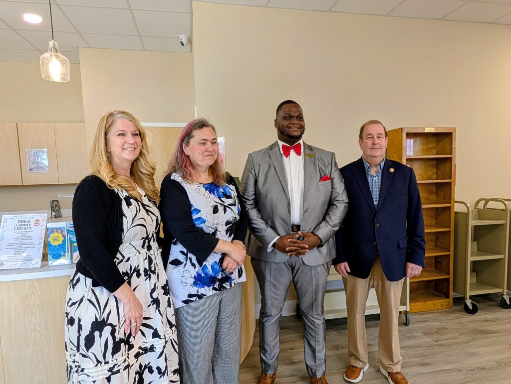 Four people standing side by side in a library, two women in dresses and two men in suits, posing and smiling at the camera.
