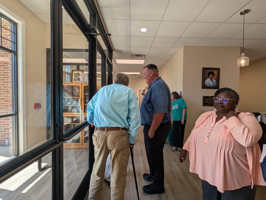 Four people stand in a hallway with glass walls and framed photos, one using a cane, while a woman in pink shirt stands in the foreground near a portrait.