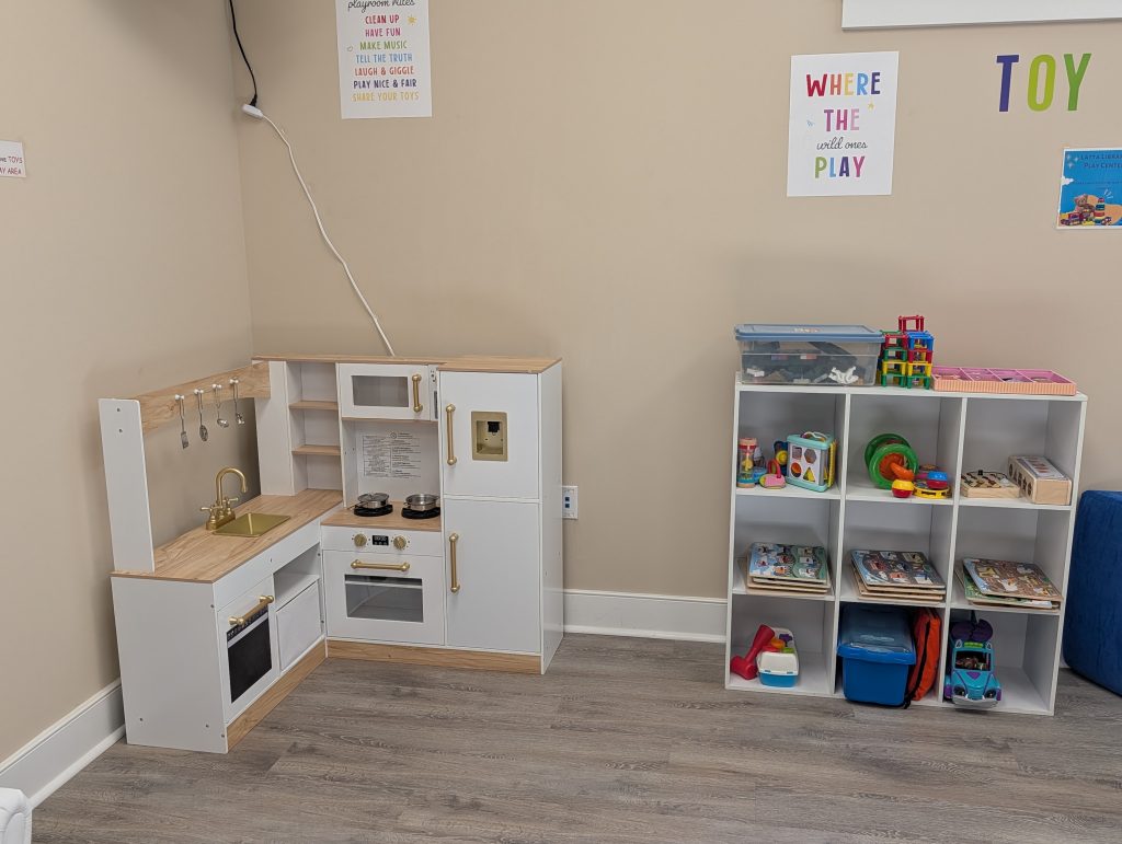 Children’s playroom with a toy kitchen, shelves organized with puzzles, bins, and colorful toys. Posters and playroom rules decorate the beige walls.