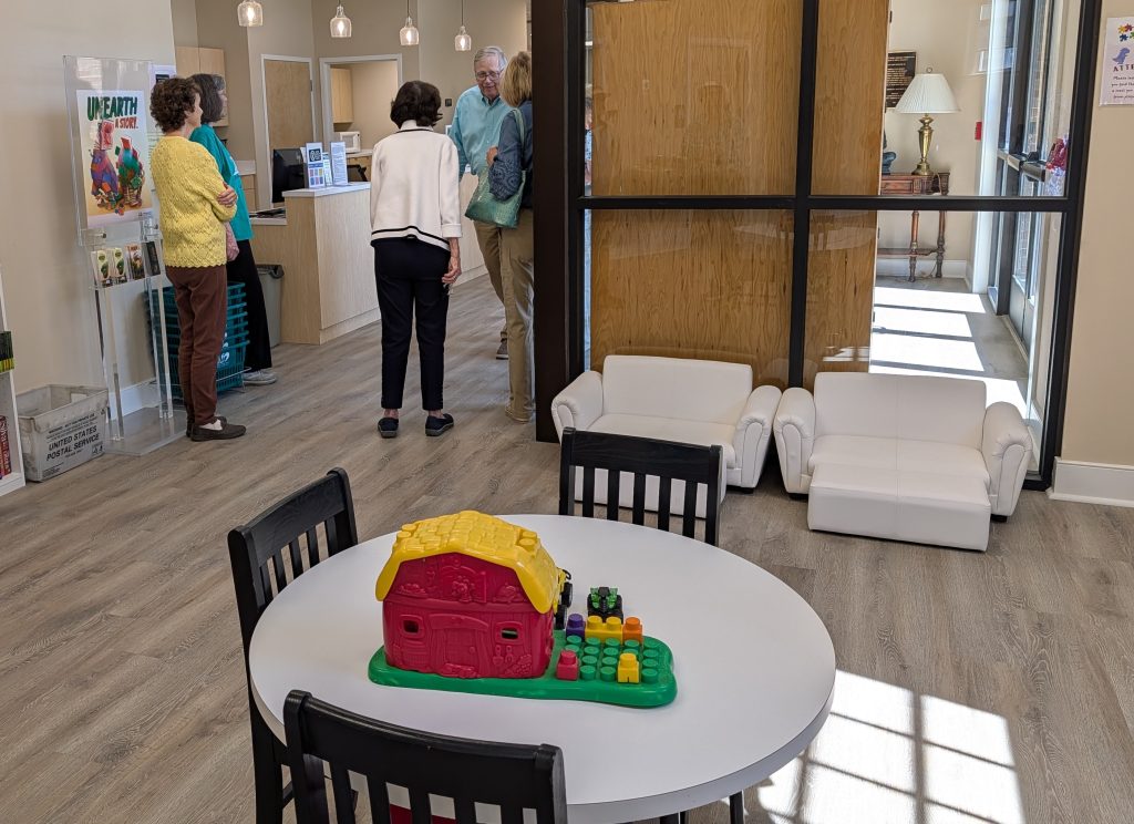 Children’s play area with a round table, toy barn, small white couches, and adults talking near a library counter in the background.