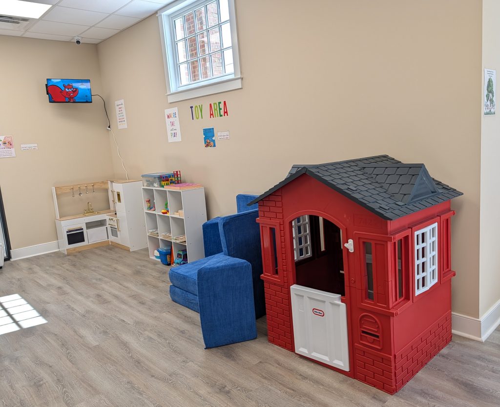 Children’s playroom with blue chairs, a red playhouse, toy kitchen, shelves with toys, TV showing cartoon, and “Toy Area” sign on beige wall.