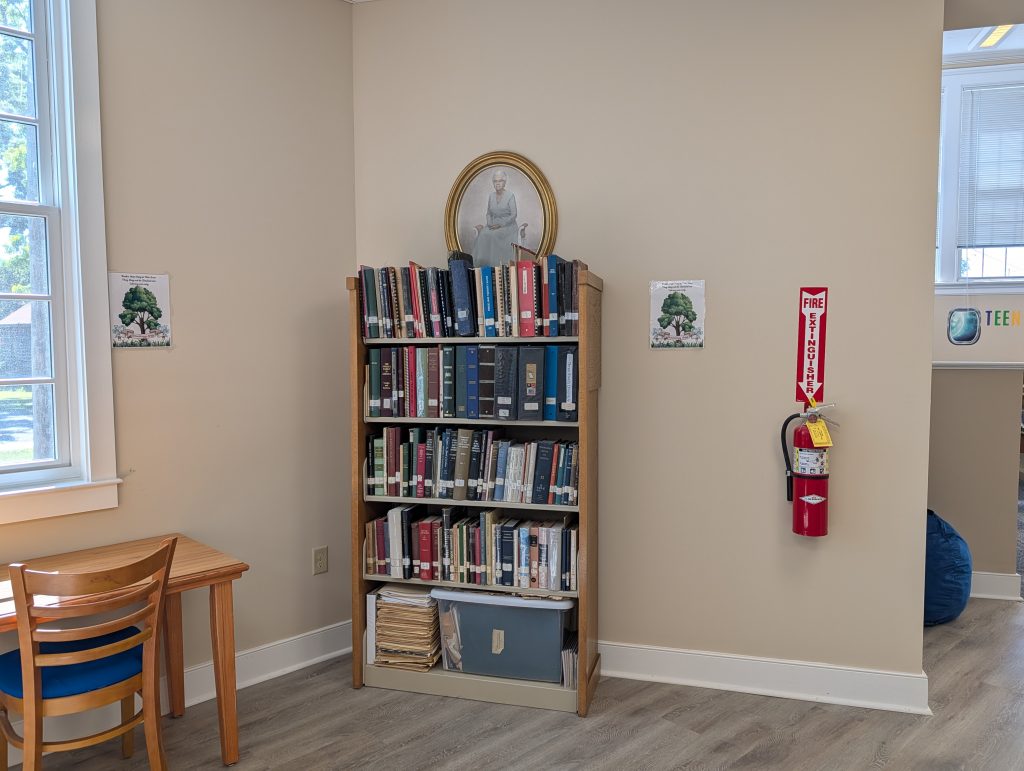 Bookshelf with books and papers, framed portrait above, fire extinguisher mounted on wall, two tree posters, wooden desk and chair to the left, window with natural light.