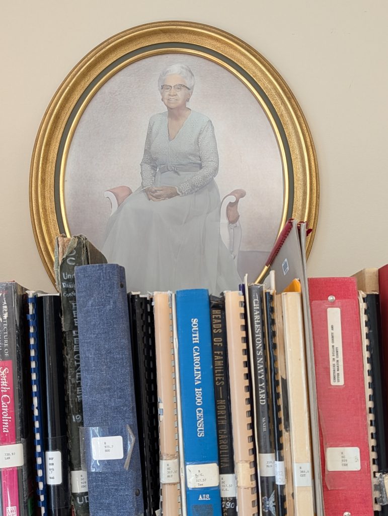 Framed portrait of elderly woman in light dress above a row of assorted books and binders, some labeled with South Carolina-related titles.