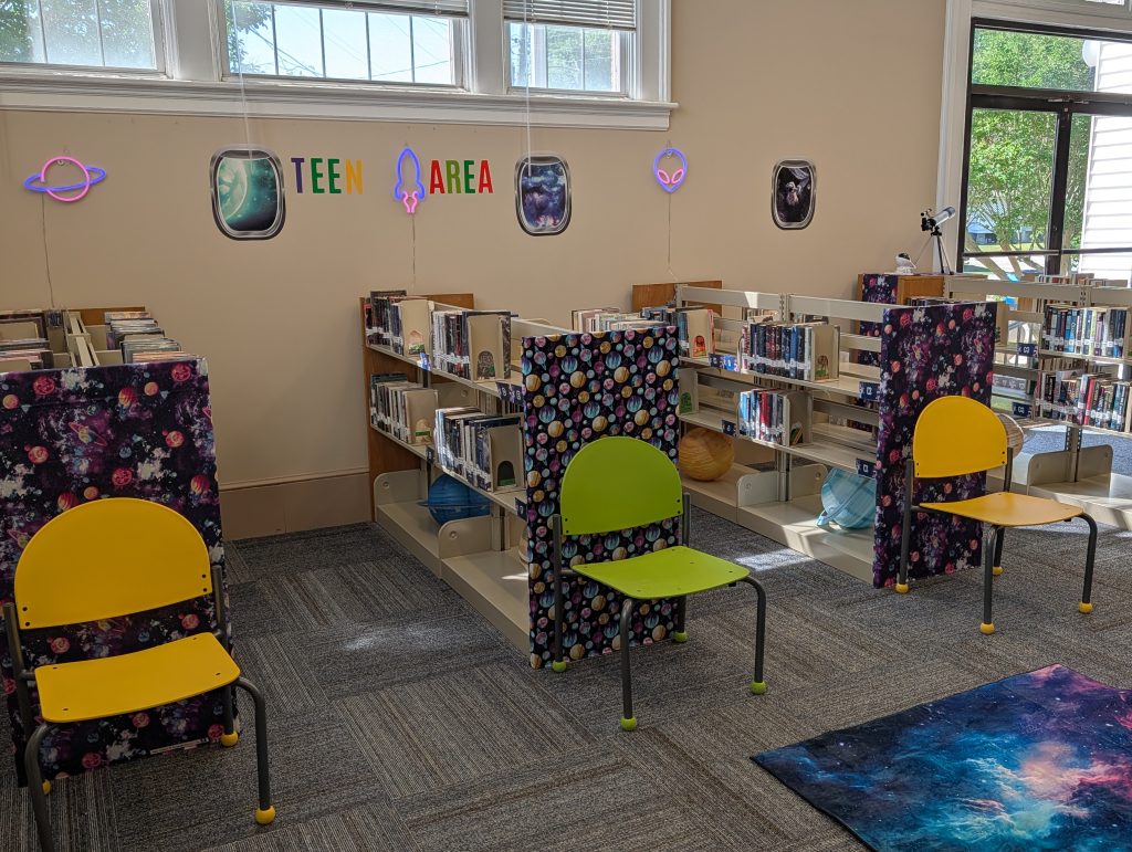 Teen library area with colorful space-themed decor, neon lights, bookshelves of young adult books, yellow and green chairs, and large windows letting in natural light.
