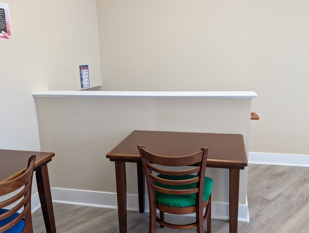 Two wooden tables with chairs, one with a green cushion, placed near a short beige wall in a simple, well-lit room with light wood flooring.