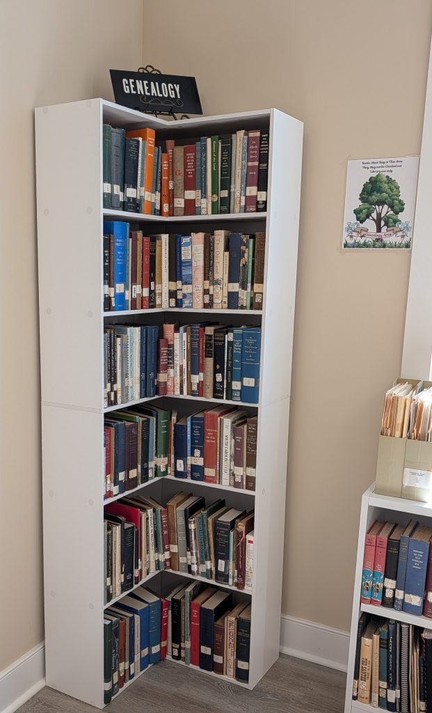 A white corner bookshelf filled with genealogy books, a "GENEALOGY" sign on top, and a small family tree-themed sign on the beige wall nearby.