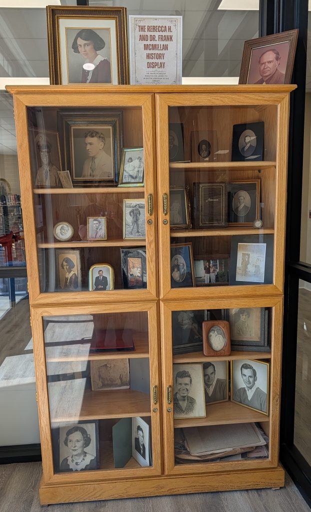 Wooden display cabinet filled with framed vintage portraits and photographs, labeled “The Rebecca H. and Dr. Frank McMillan History Display” on top.