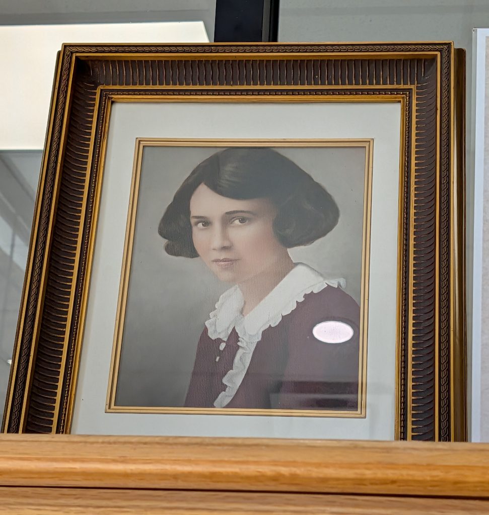 Portrait of a woman with short, dark hair in a vintage frame, wearing a maroon dress with a white ruffled collar, displayed on a shelf.