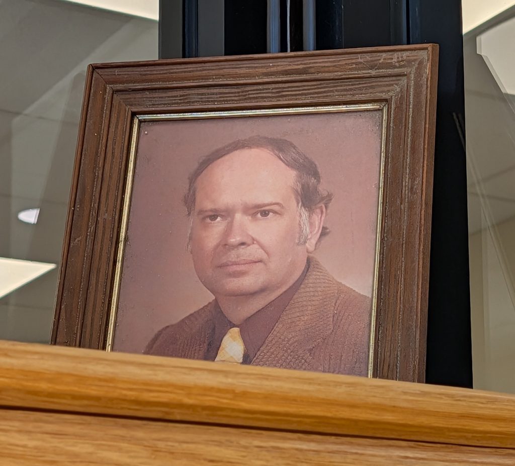 A framed vintage portrait of a middle-aged man in a brown suit and yellow tie, displayed on a wooden shelf.