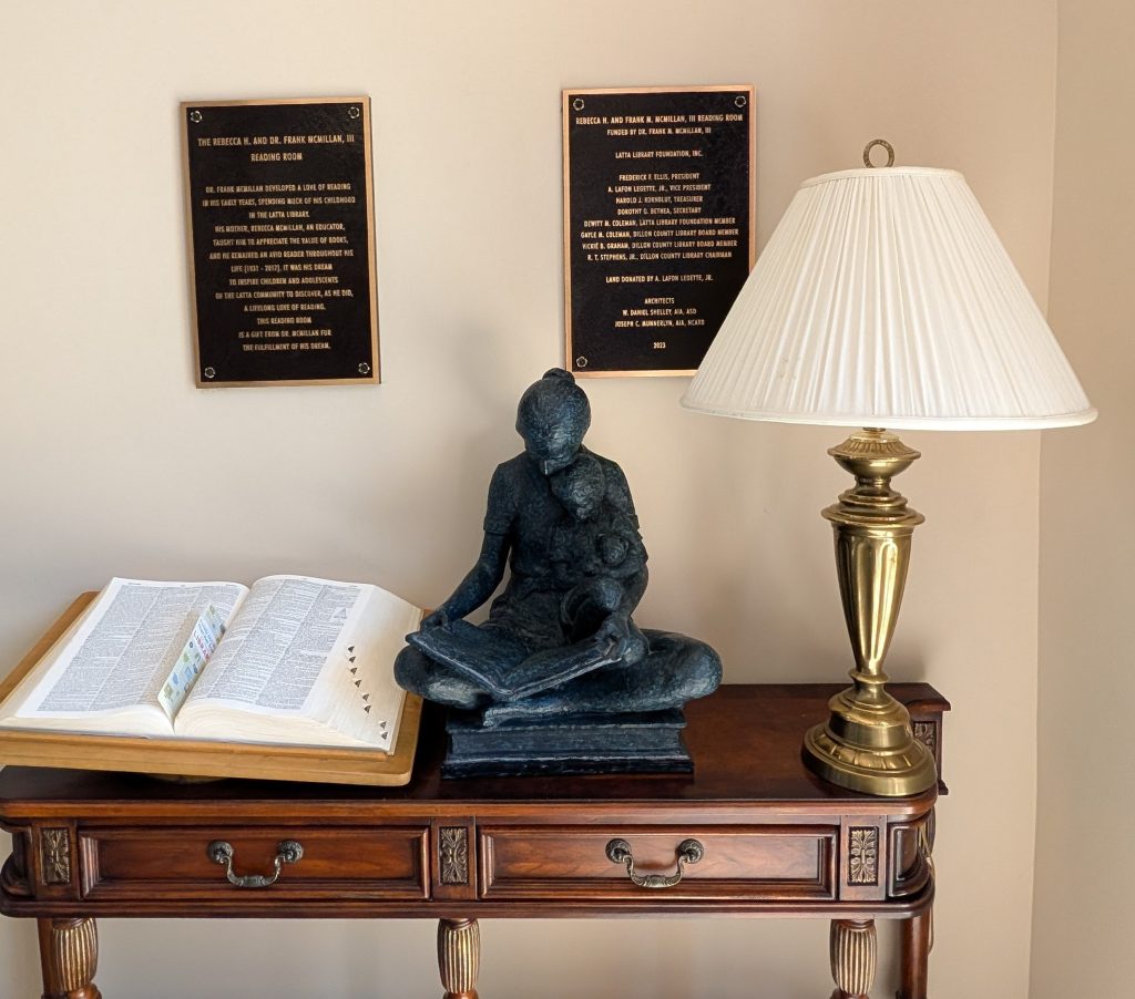 A wooden table with an open book, bronze statue of a person reading to a child, a brass lamp, and two commemorative plaques on the wall.