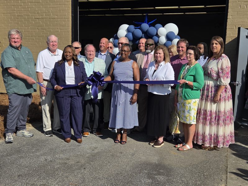 A group of people gathers for a ribbon-cutting ceremony outside a building, with blue and white balloons visible in the background.