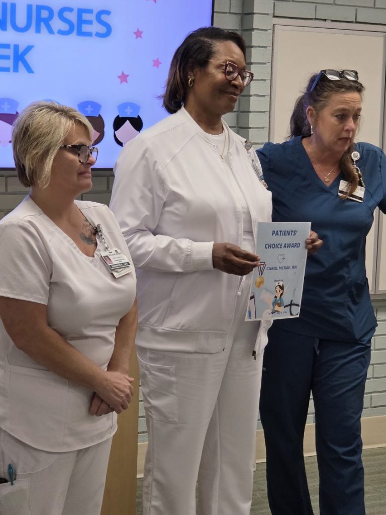 Three nurses standing together; one is holding a "Patients' Choice Award" certificate. A "Nurses Week" sign is visible in the background.