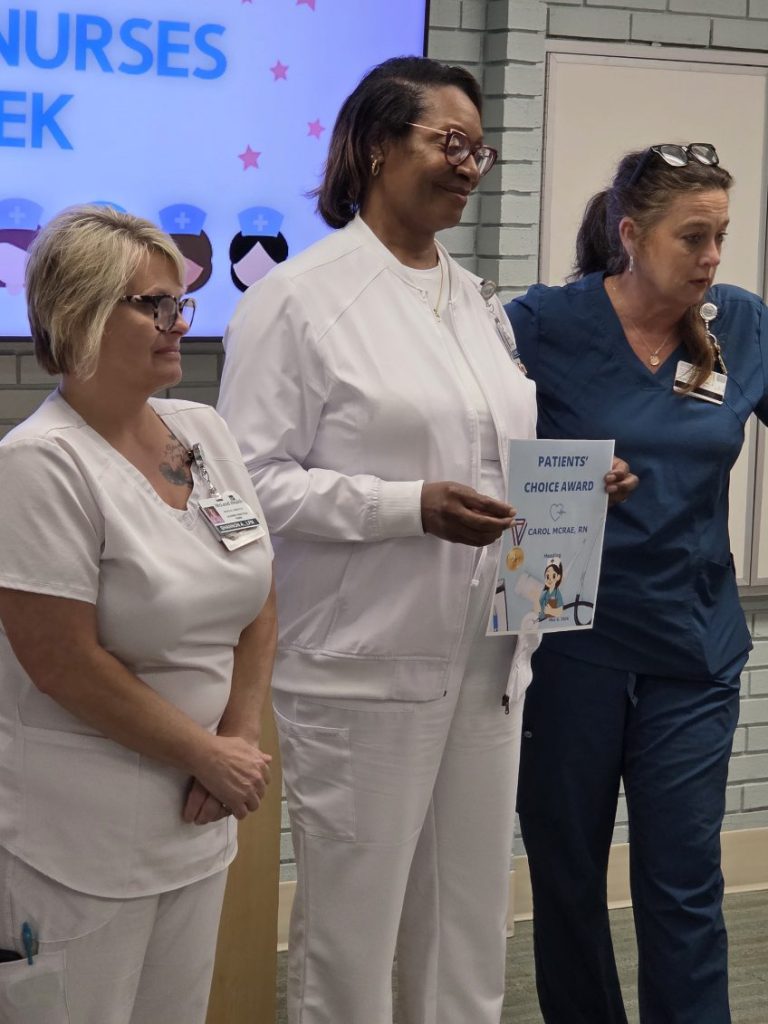 Three nurses stand together, one holding a "Patients' Choice Award" certificate. A sign in the background reads "Nurses Week" with cartoon nurse graphics.