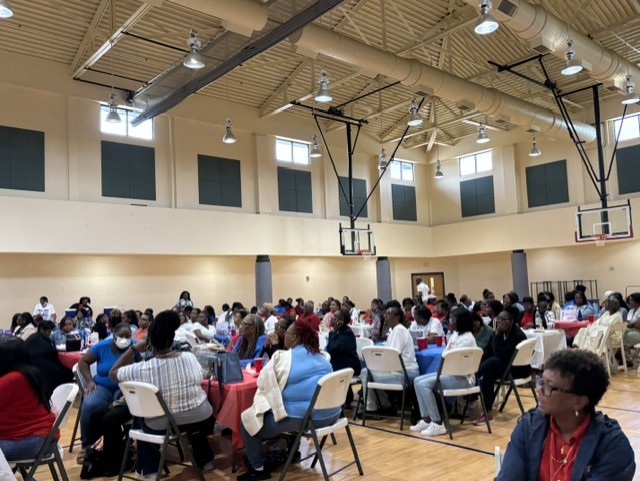 Large group of people seated at tables in a gymnasium, attending an indoor event or gathering with high ceilings and bright lighting.