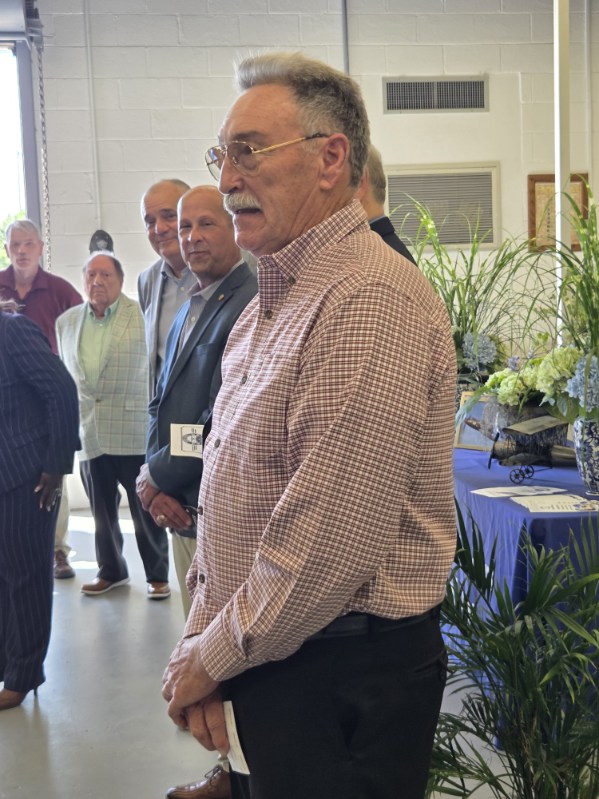 A man in a checkered shirt stands in the foreground with a group of older men behind him, indoors with plants and a table on the side.
