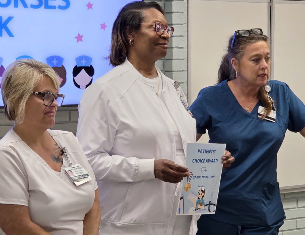 Three nurses standing together; one holds a "Patients' Choice Award" certificate. Background features a screen with nurse icons and partial text.