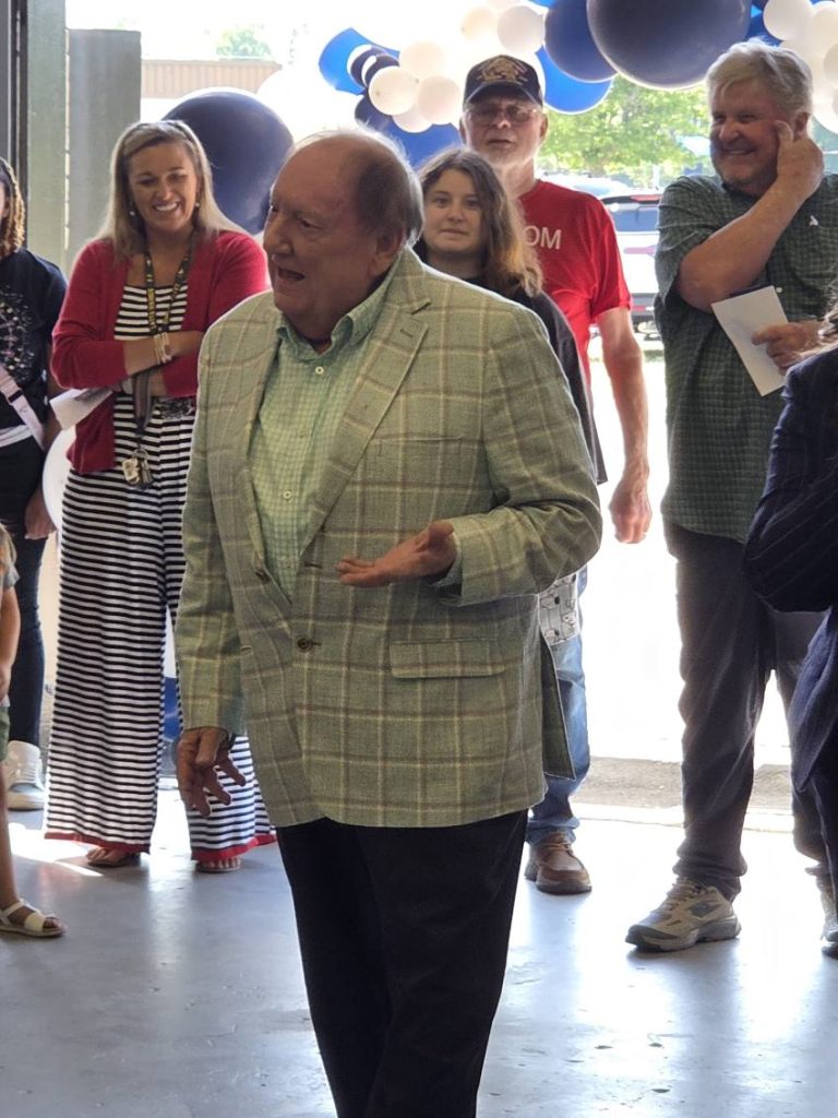 older man in a plaid blazer speaks to a group of people indoors, with blue and white balloons in the background.