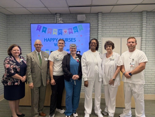 A group of seven people, including nurses in uniforms, stand together smiling in front of a "Happy Nurses Week" sign.