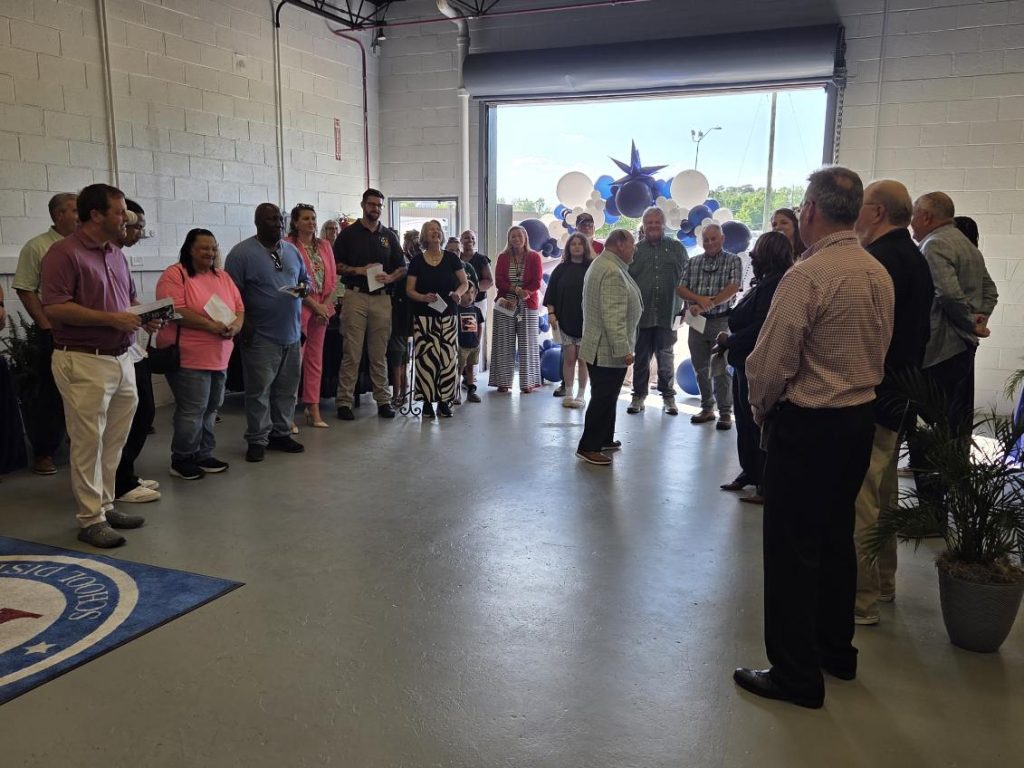 A group of people standing in a semi-circle indoors, listening to a speaker. Balloon decorations are visible by a large open door in the background.