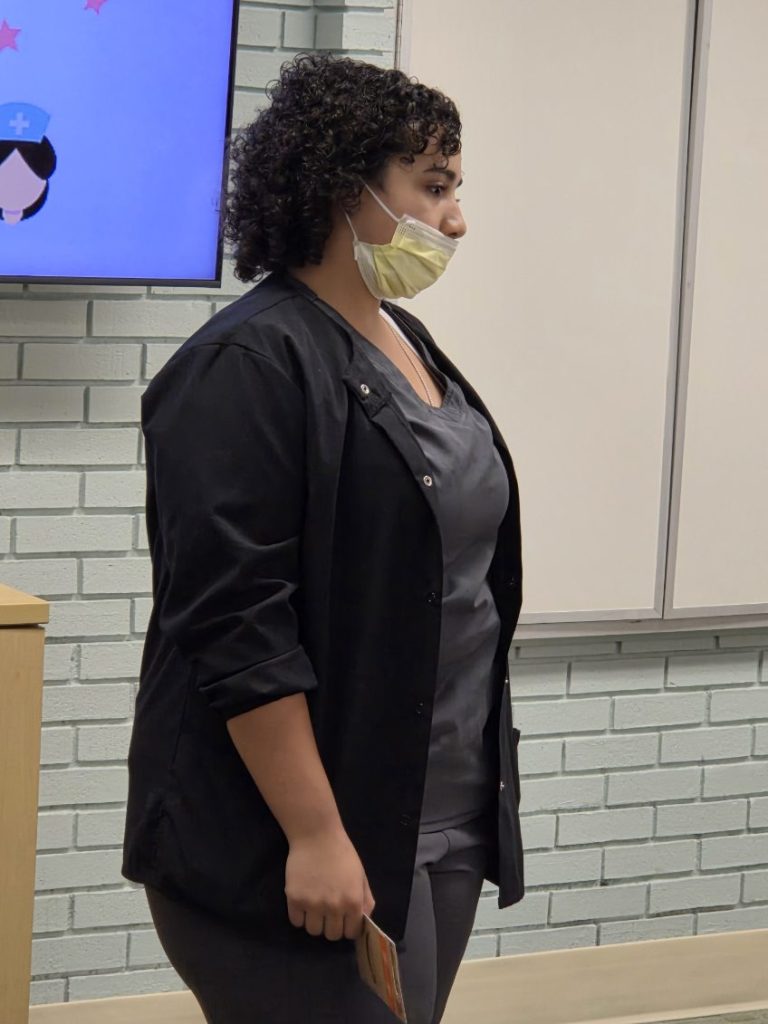 Woman in scrubs and face mask standing indoors near a whiteboard, holding a booklet, with a screen showing a nurse cartoon in the background.