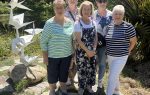 Five women stand outdoors on a sunny day near a metal crane sculpture and greenery, smiling for the photo.