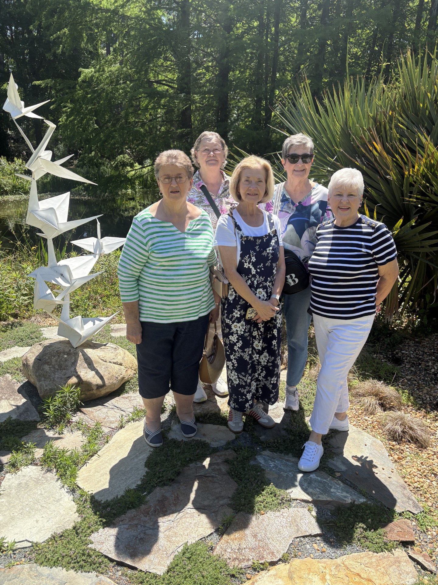 Five women stand outdoors on a sunny day near a metal crane sculpture and greenery, smiling for the photo.