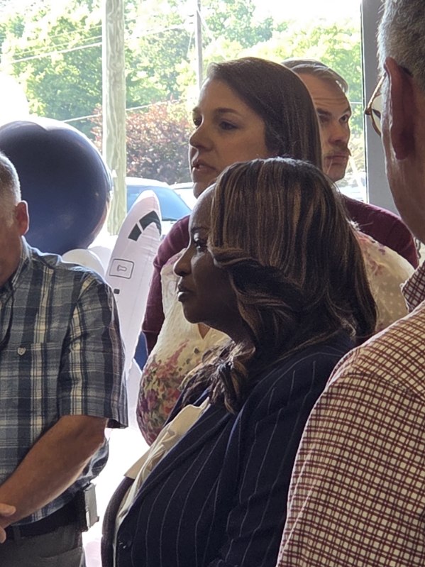 Group of adults standing indoors, attentively listening. Background shows windows with trees and a balloon. People are dressed in business casual and daylight is coming through the glass.