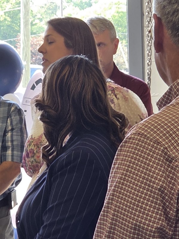 A group of people standing indoors, some facing the camera and others with their backs turned, engaged in conversation near a window.