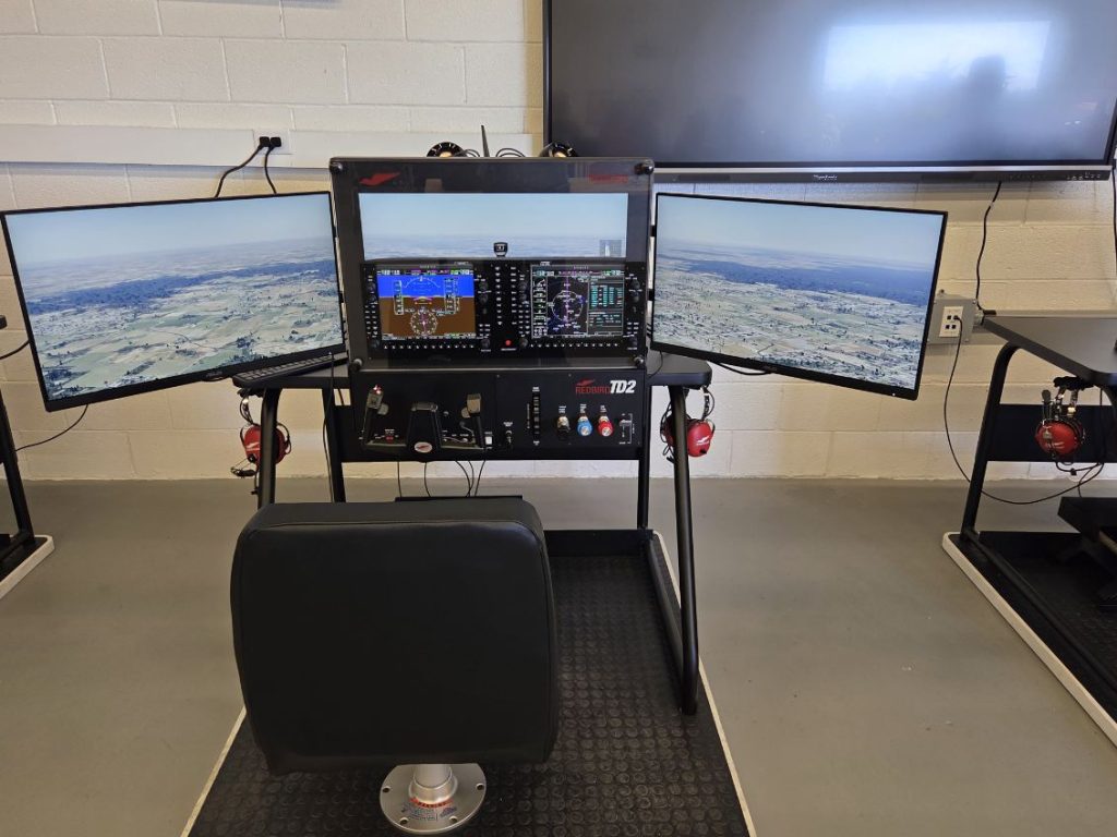 A flight simulator with three monitors displaying aerial landscapes, flight controls, and a black chair positioned in front in a classroom setting.