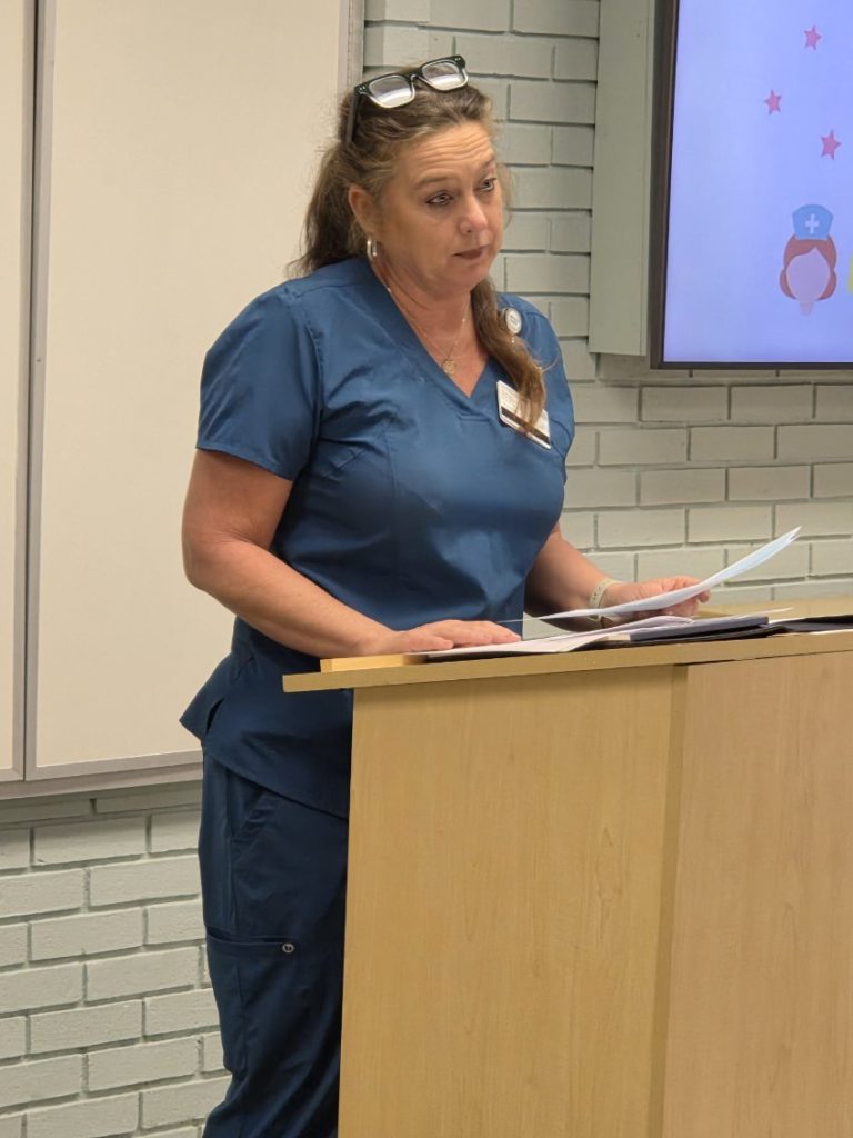 A woman in blue scrubs stands at a podium, holding papers and speaking, with a presentation screen and white brick wall in the background.