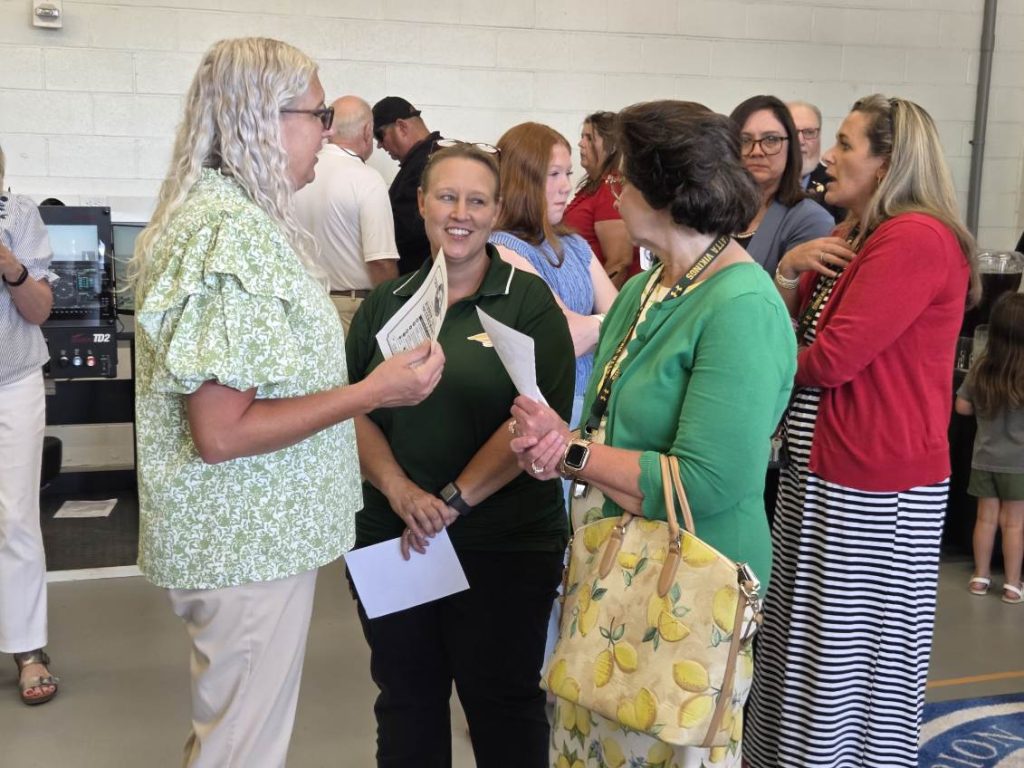 A group of adults, mostly women, stand and talk in a casual indoor setting. Some hold papers, and one person carries a lemon-patterned handbag.