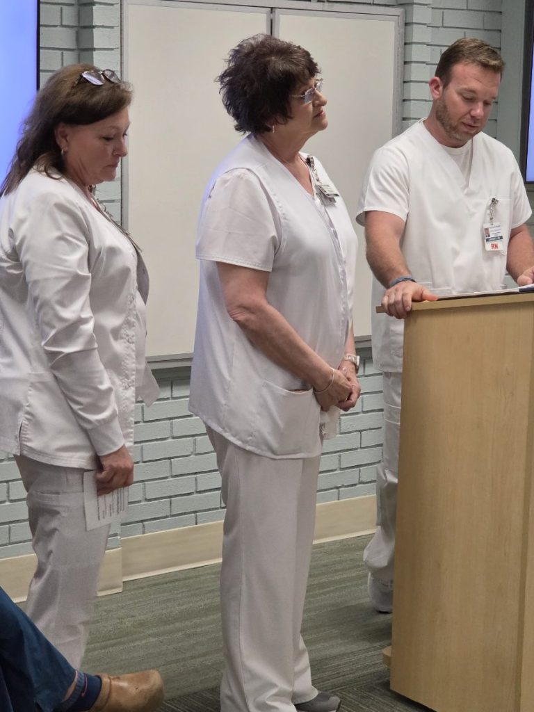 Three nurses in white uniforms standing indoors, one reading from a podium, appearing to participate in a meeting or presentation.