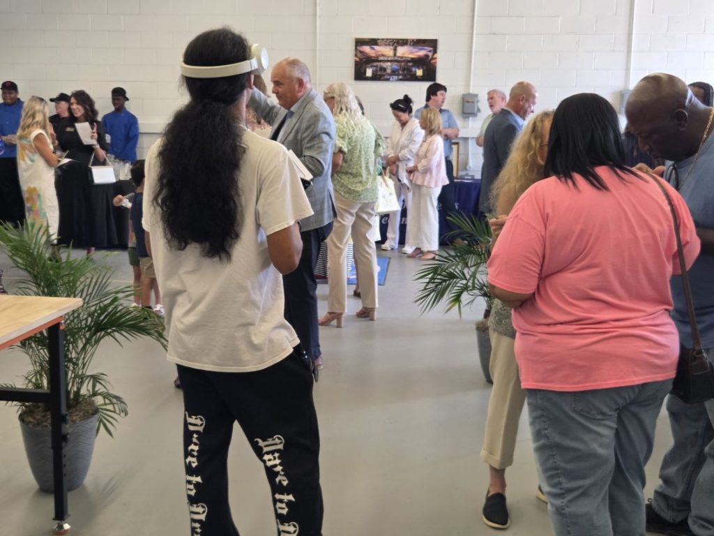 People gather in a spacious indoor venue, chatting in groups near potted plants; some stand in line at a counter in the background.