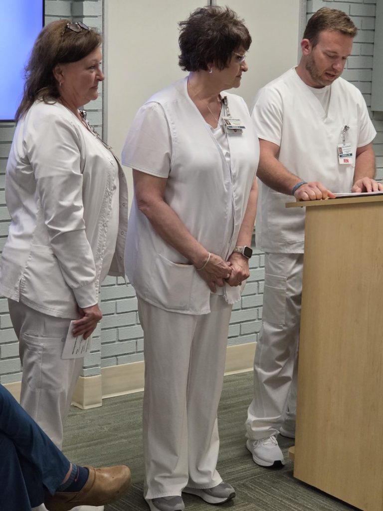 Three nurses in white uniforms stand together, with one reading from papers at a podium in a clinical or hospital setting.