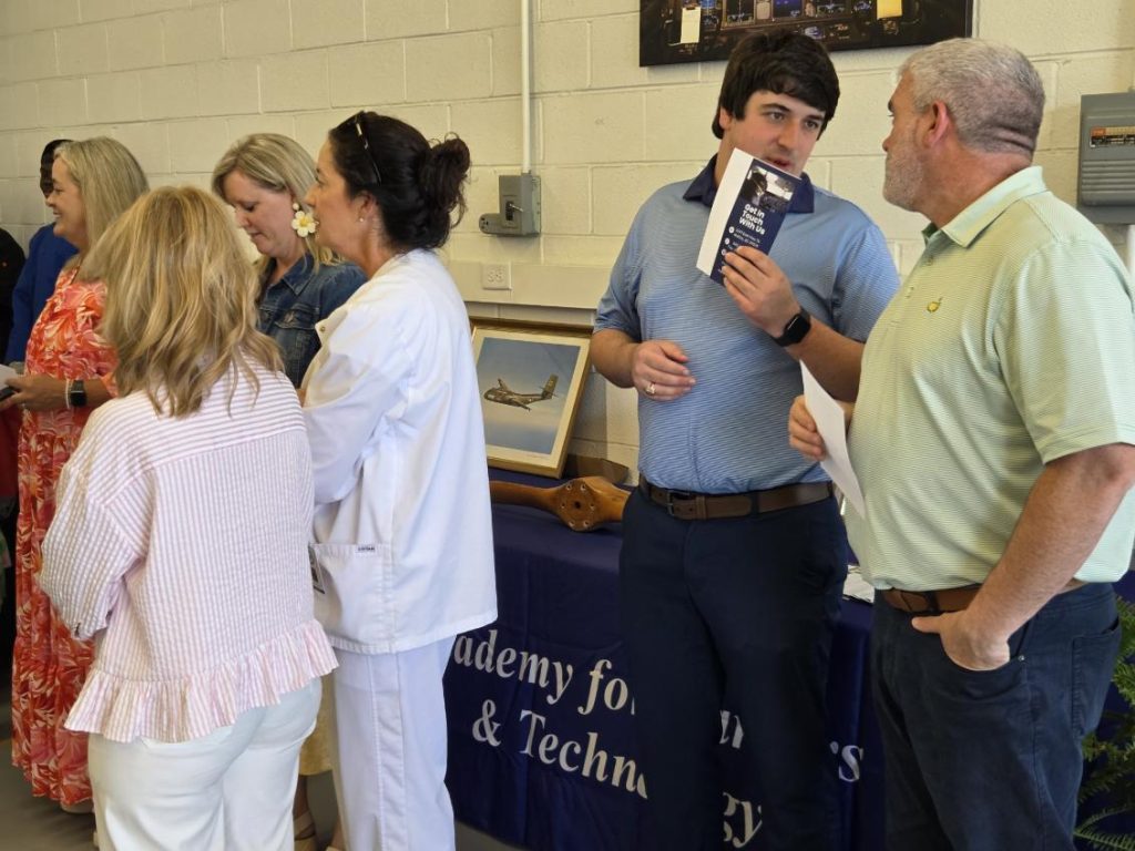 Several people stand and converse indoors near a table with aviation photos and brochures, in a room with white brick walls and informational displays.