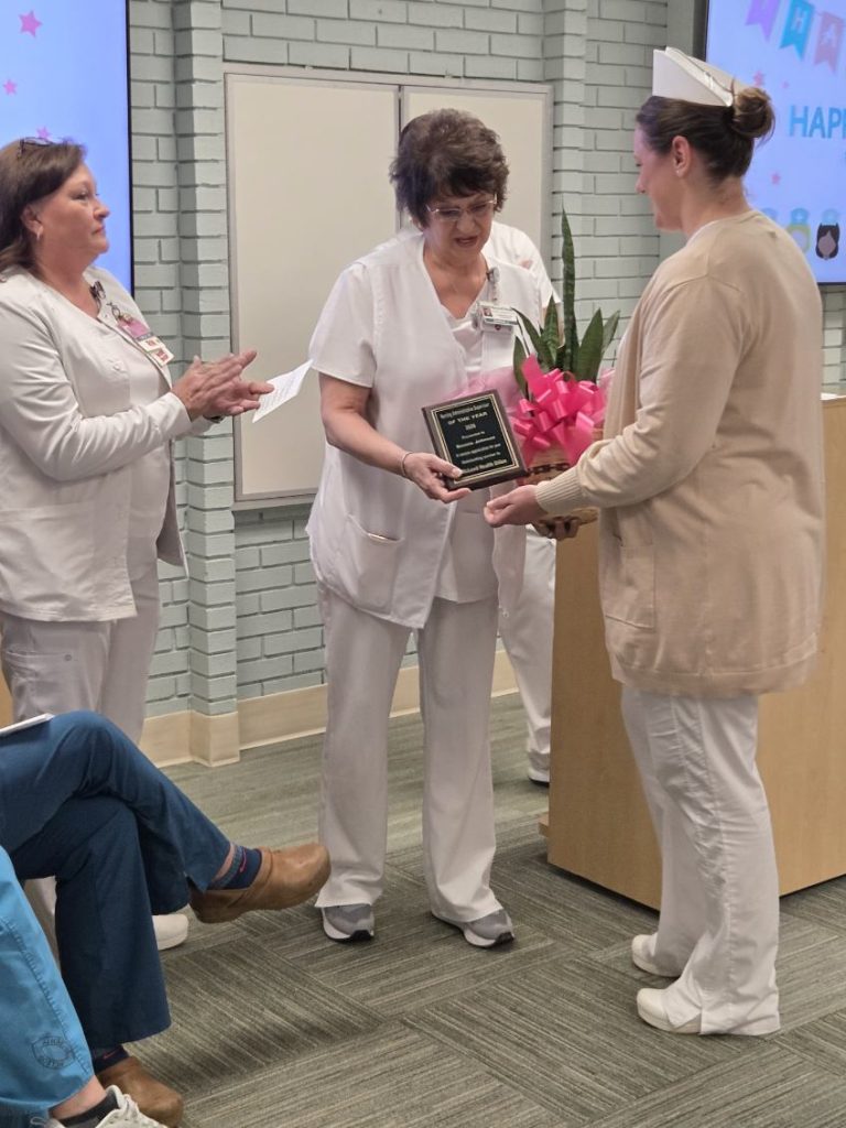 A nurse receives an award plaque and a plant with a pink ribbon from colleagues in a ceremony at a healthcare facility.