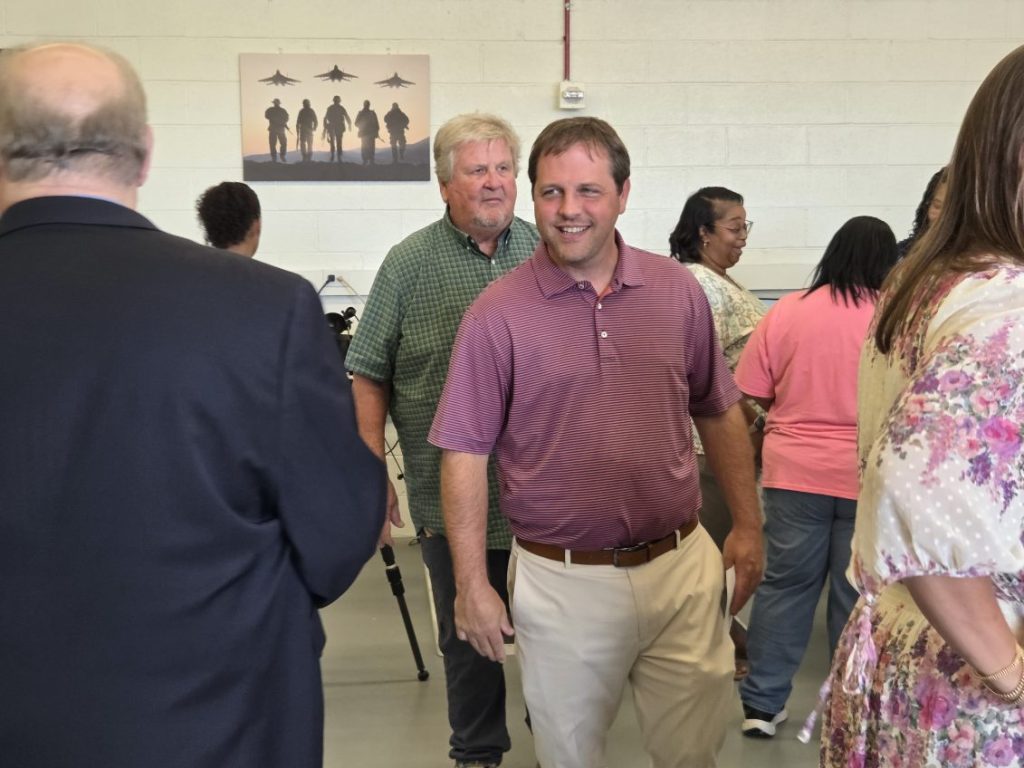 People socializing indoors; a man in a maroon shirt smiles in the center. Military-themed artwork with jet silhouettes and soldiers hangs on the wall behind them.