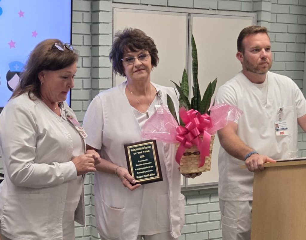 Three nurses in white uniforms; one holds a plaque and a potted plant wrapped with a pink ribbon, standing together during an award presentation in a hospital setting.