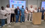 Group of nurses and staff stand in front of "Happy Nurses Week" sign, smiling during a celebration in a medical facility.