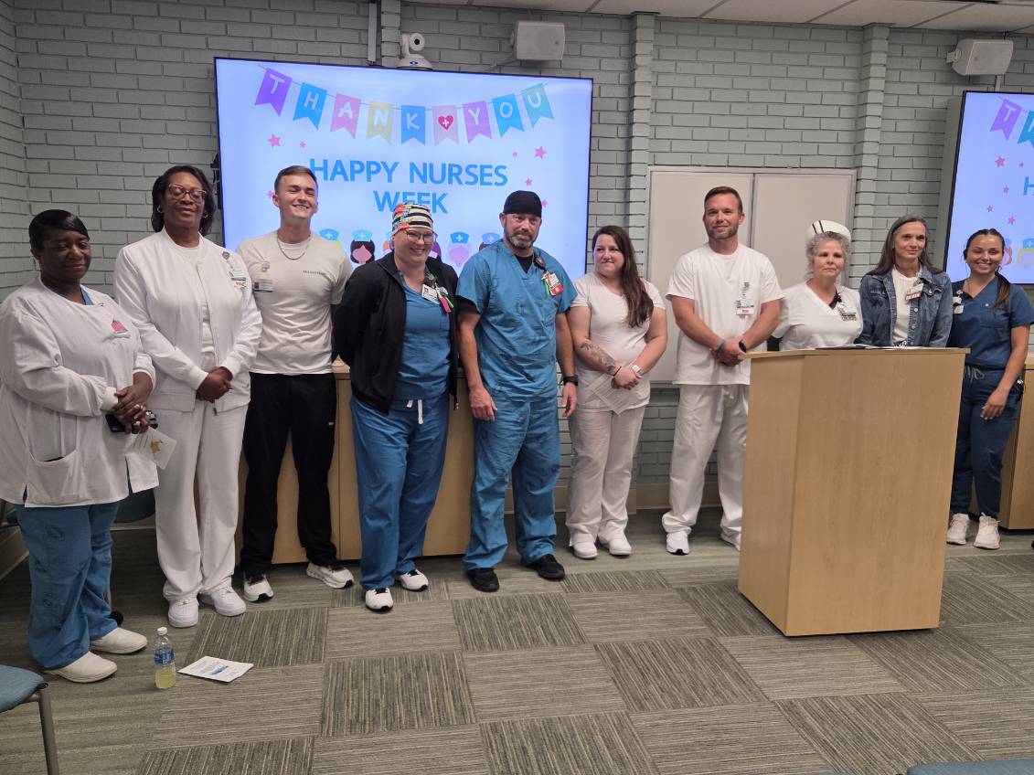 Group of nurses and staff stand in front of "Happy Nurses Week" sign, smiling during a celebration in a medical facility.
