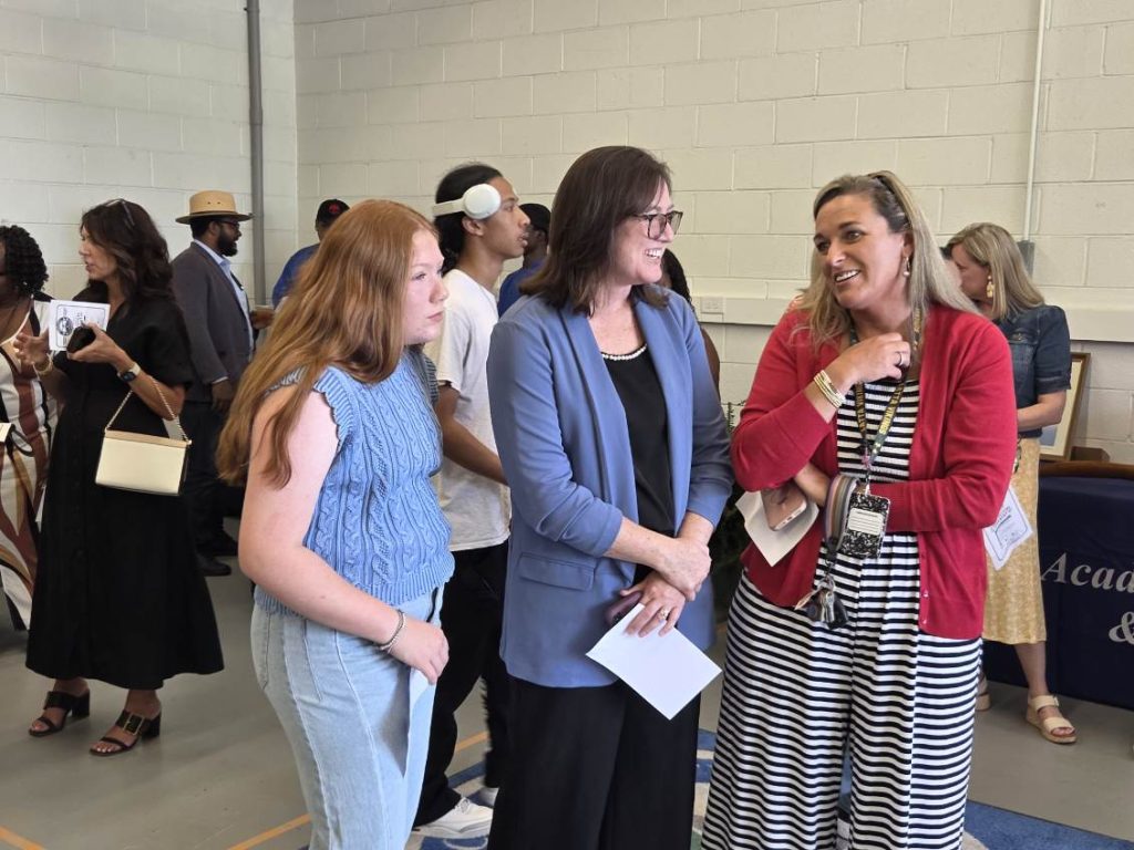 A group of people, mostly women, stand indoors having conversations at an event. Three women are talking and smiling in the foreground.