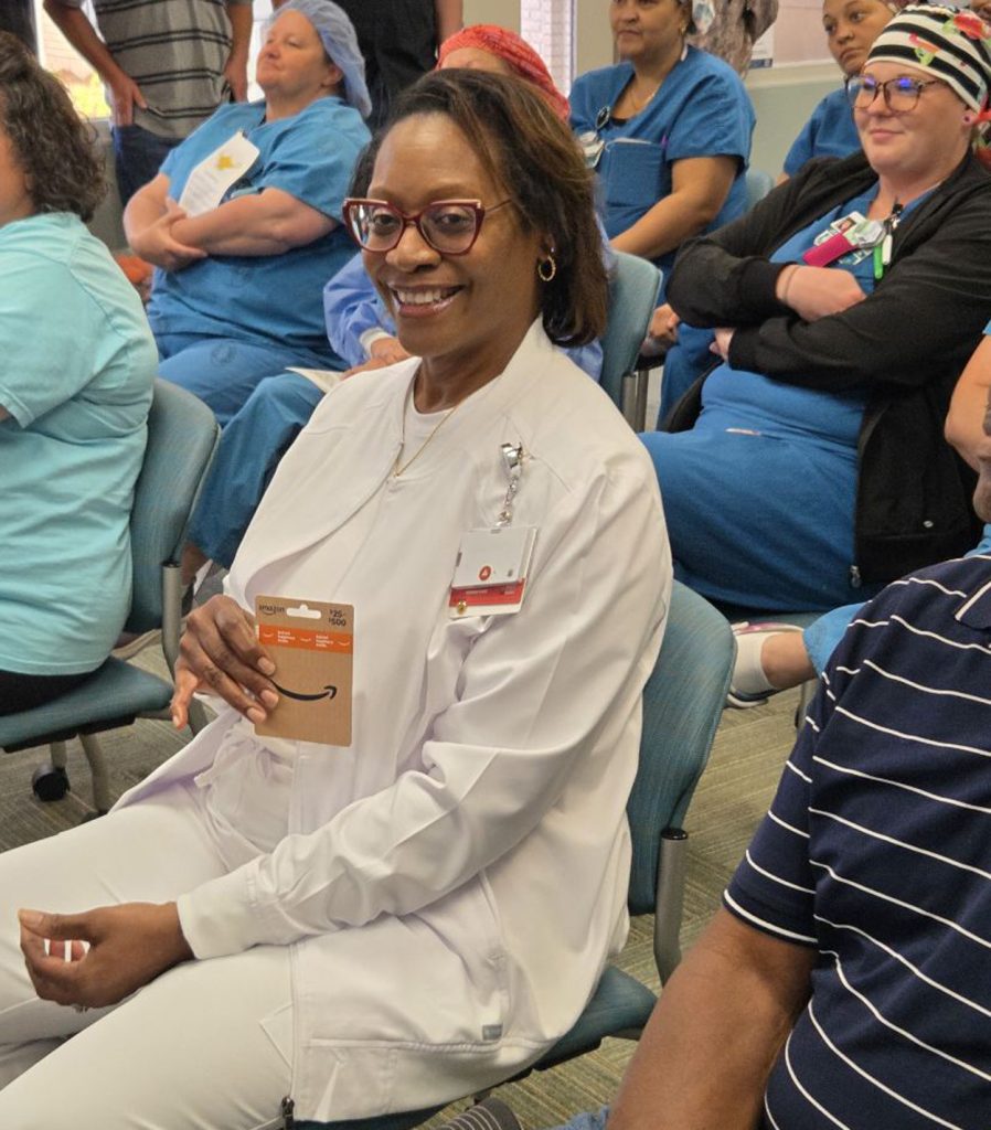 Smiling healthcare worker in white holding an Amazon gift card, sitting among colleagues in blue scrubs during what appears to be an award or recognition event.