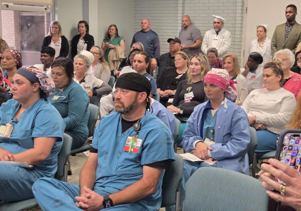 A group of healthcare workers and staff, some in scrubs and uniforms, attentively listen during a meeting or event in a hospital or medical facility.
